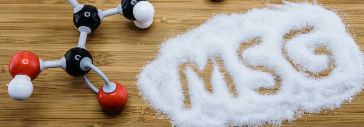 A ball-and-stick glutamate molecular model on the left and white MSG crystals spelling the word MSG on a wooden surface on the right