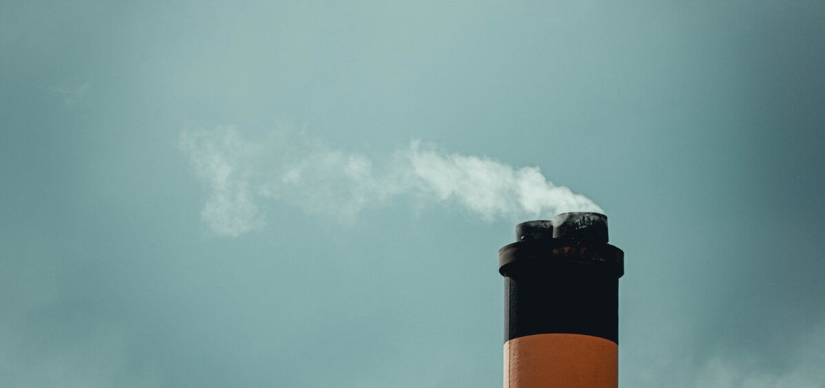 Industrial smokestack with a rust-orange and black exterior emitting a white steam plume against an overcast grey sky — stack emission source treated by activated carbon odor control