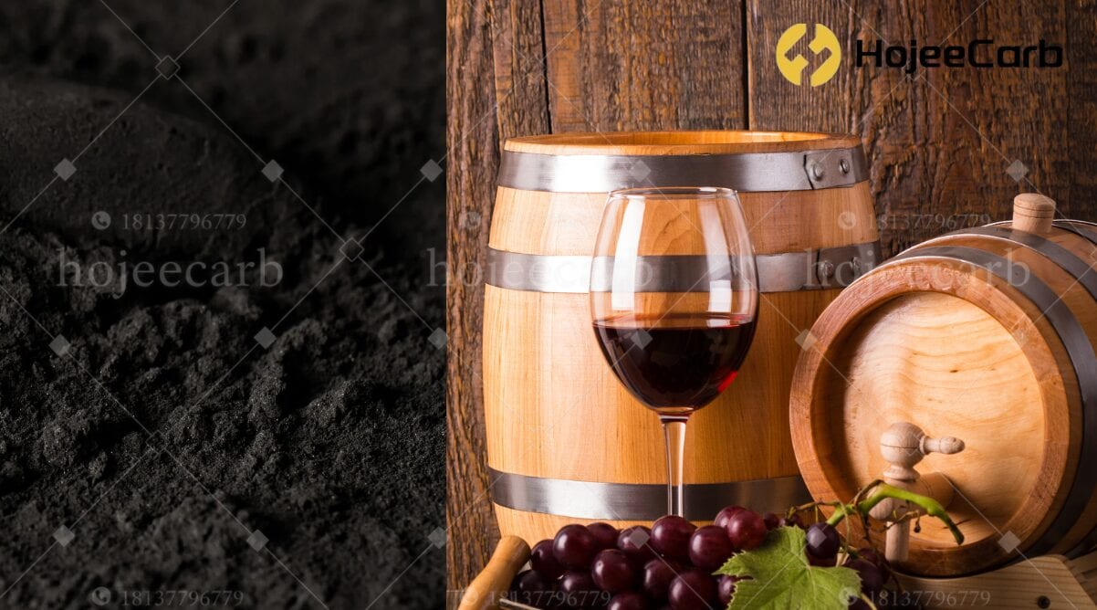 Black activated carbon granules filling the left half; a glass of red wine beside a large upright oak barrel with steel hoops and a small horizontal barrel with a wooden spigot on a rustic wood-plank surface on the right — HojeeCarb wine-grade carbon