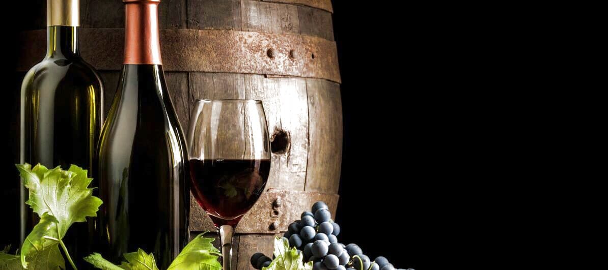 Two unlabeled wine bottles, a glass of deep red wine, a wooden oak aging barrel and a cluster of dark blue-purple grapes with green vine leaves